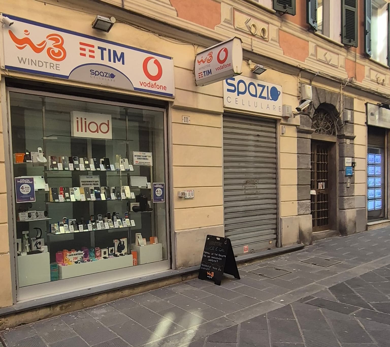 A small phone shop on an Italian pedestrian street, its sign showing the WindTre, TIM, and Vodafone logos above a window of phones for sale.