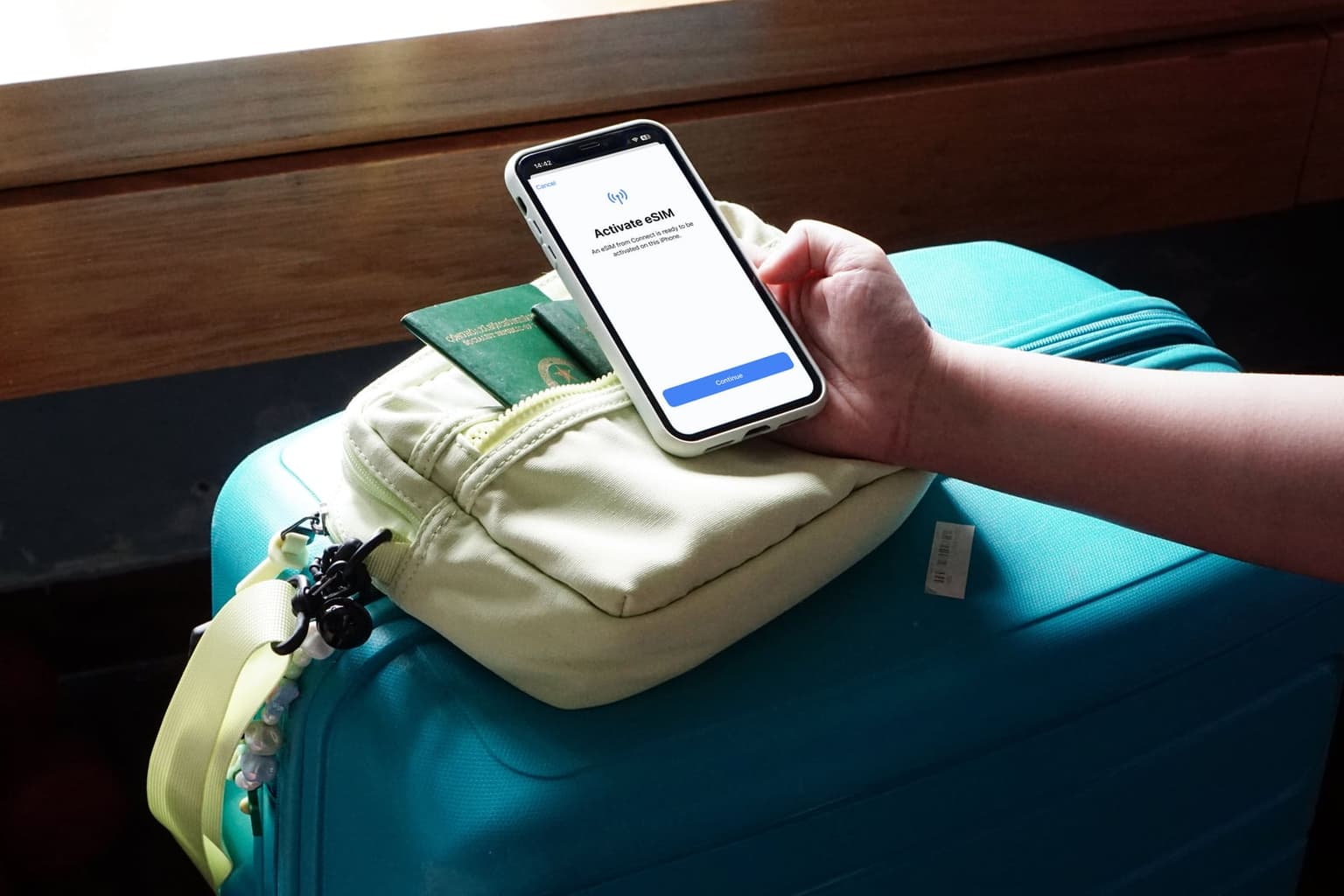 A hand holds an iPhone showing the Activate eSIM screen above a packed shoulder bag and a teal carry-on, a passport tucked into the bag's pocket.