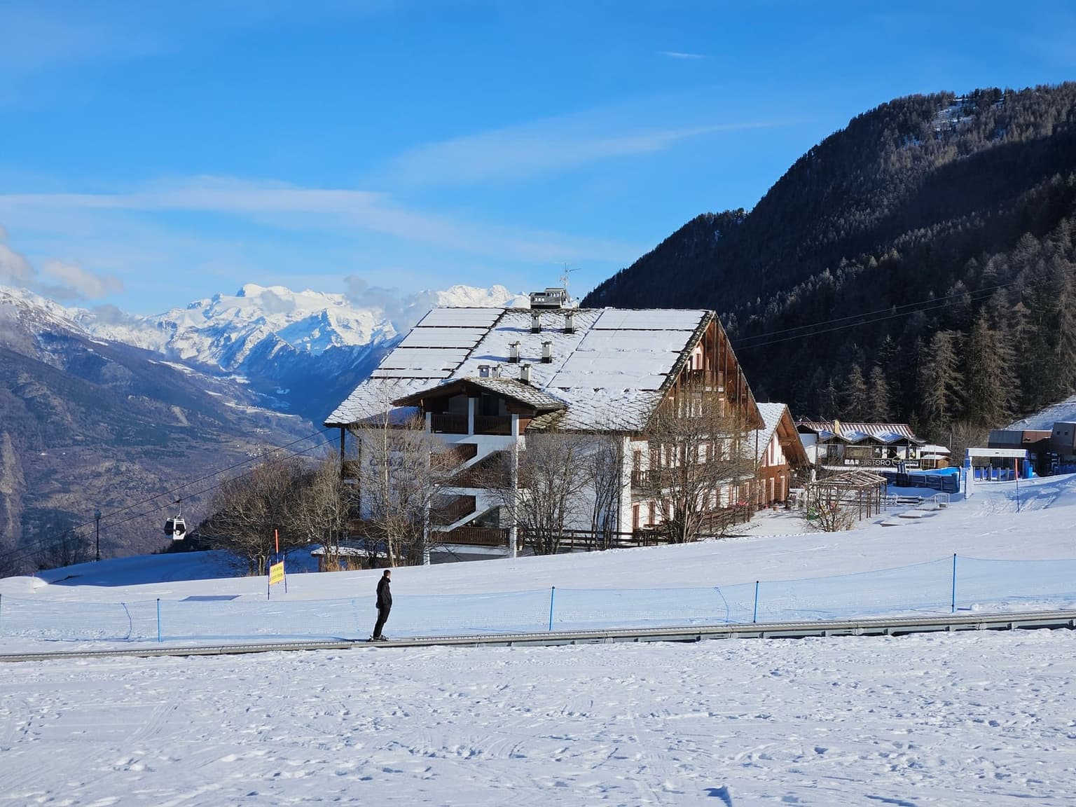 A lone skier riding a moving-carpet lift across a gentle beginner area beside a large snow-roofed chalet, with a gondola cabin crossing toward a deep valley and a high snowy massif on the horizon