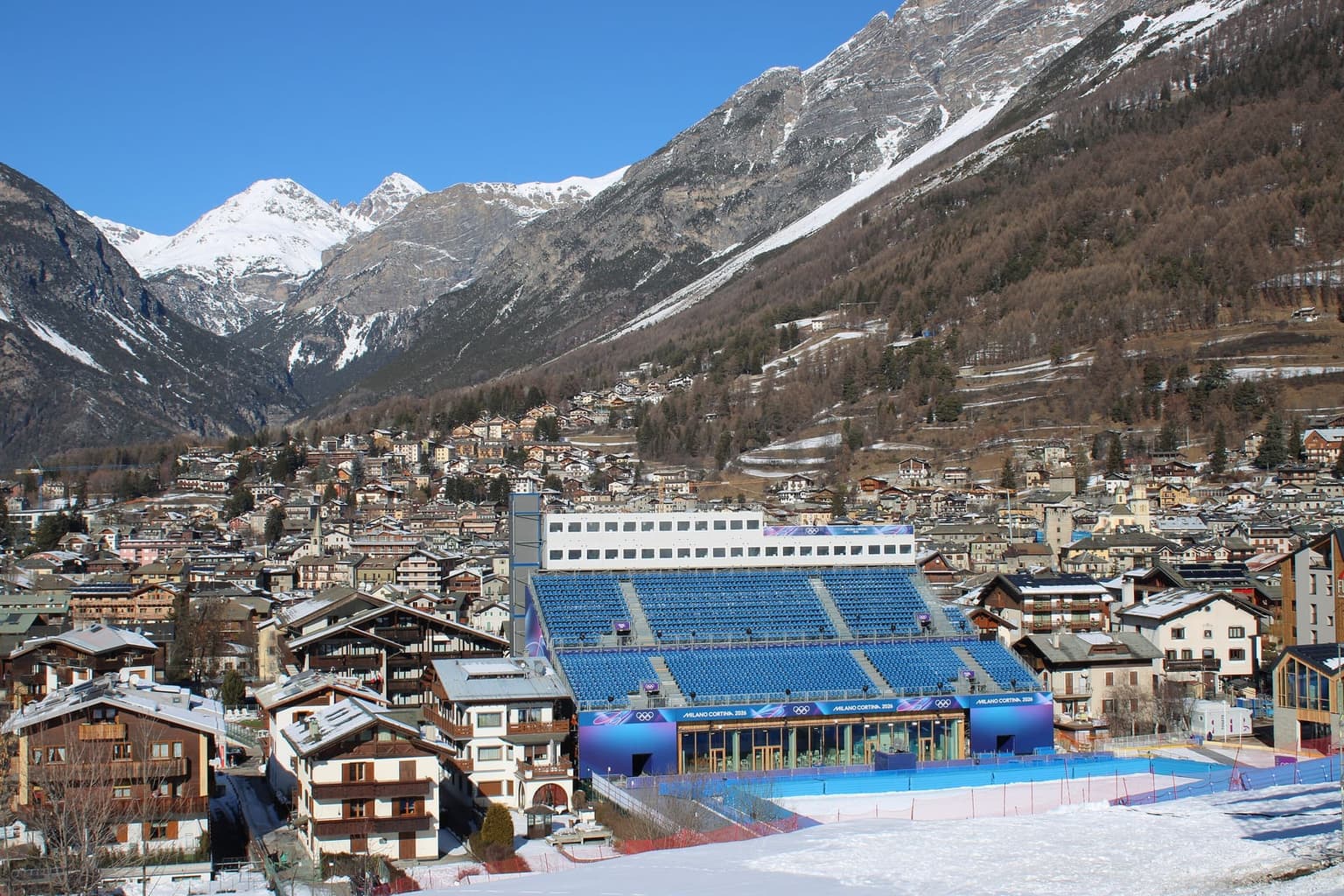 Bormio's rooftops filling the valley below steep mountain slopes, with the blue-seated Milano Cortina 2026 grandstand at the snowy finish area of the Stelvio ski course