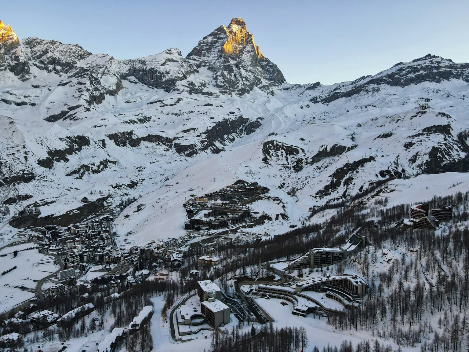 The Matterhorn's Italian face catching first light above Breuil-Cervinia, with the village, ski lifts and broad snowy pistes in shade below and curved apartment buildings among the larches in the foreground