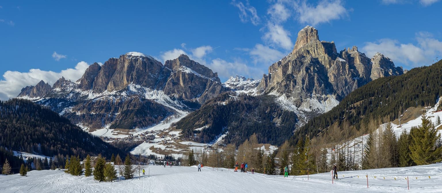 Corvara in Alta Badia in winter, the village spread across the valley floor beneath the tall rock tower of the Sassongher, with skiers on a gentle piste in the foreground