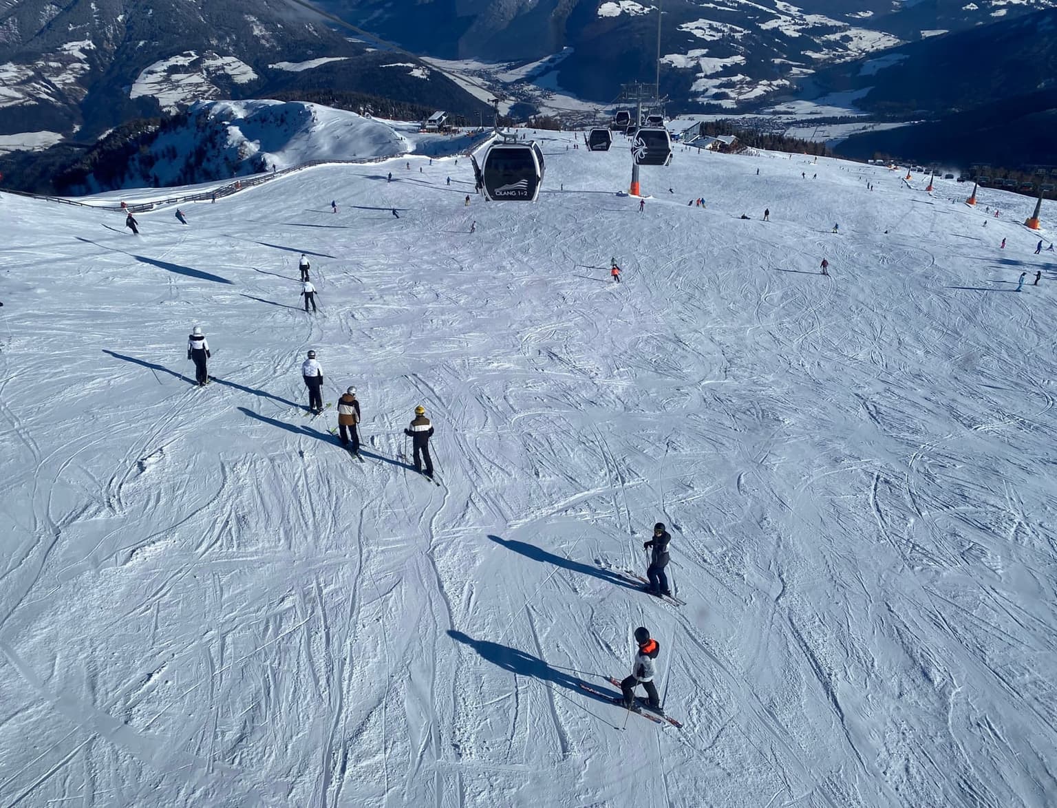 Skiers spread across a broad, sunny upper slope on Kronplatz, seen from above, with gondola cabins heading down toward the Puster Valley and its villages far below