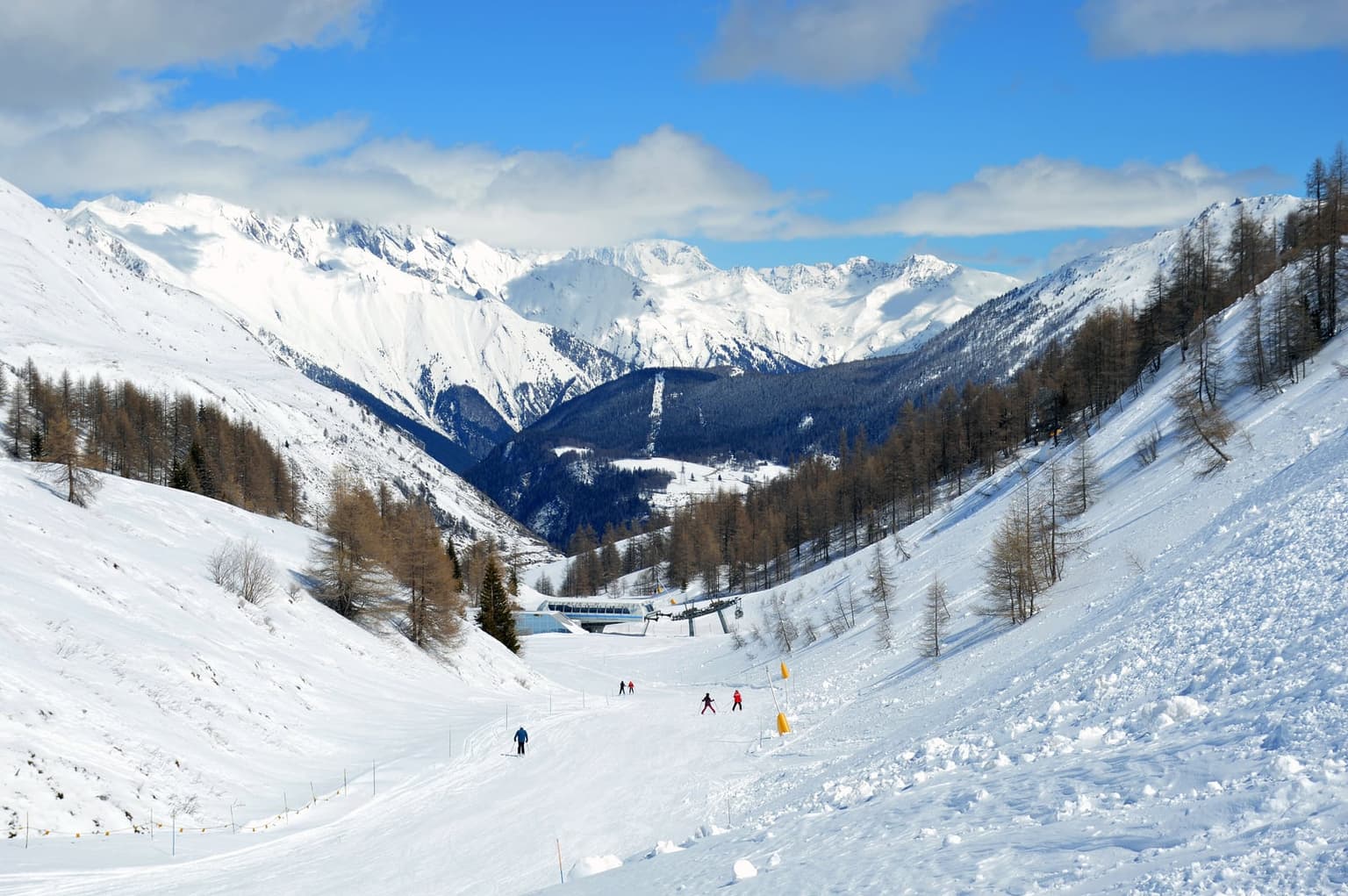 Skiers descending a wide piste through larch trees toward a chairlift base station at La Thuile, with snow-covered peaks rising across the valley under a blue sky