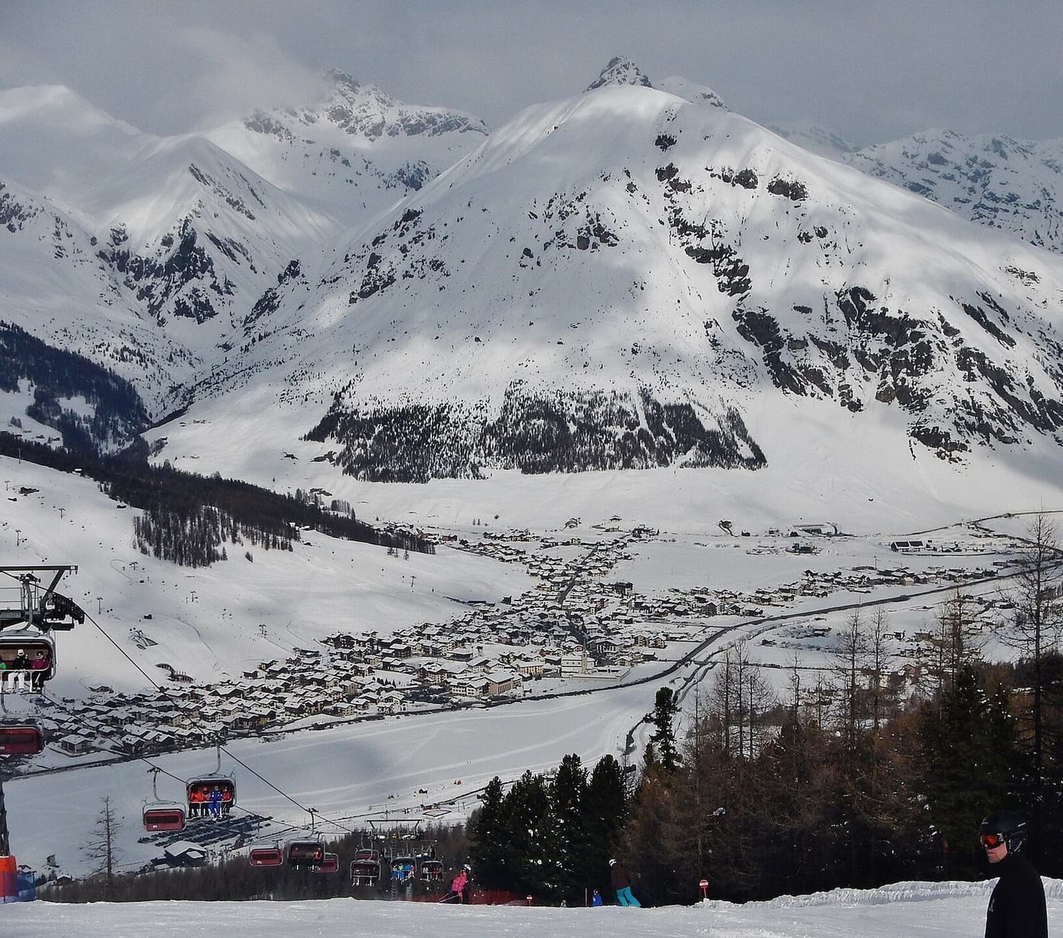 The long town of Livigno stretched along a wide, flat, snowy valley, seen from the opposite ski slope with a chairlift in the foreground and big snow-covered mountains rising behind