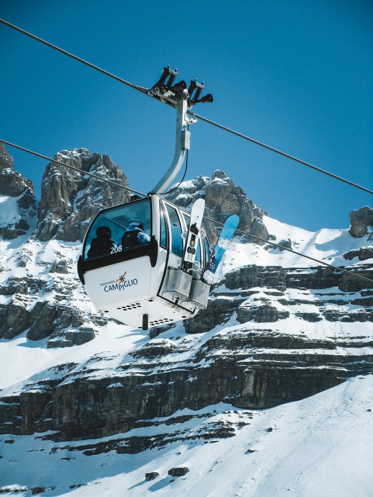 A Madonna di Campiglio gondola cabin with snowboards in its outside rack riding past the layered, snow-dusted limestone cliffs of the Brenta Dolomites