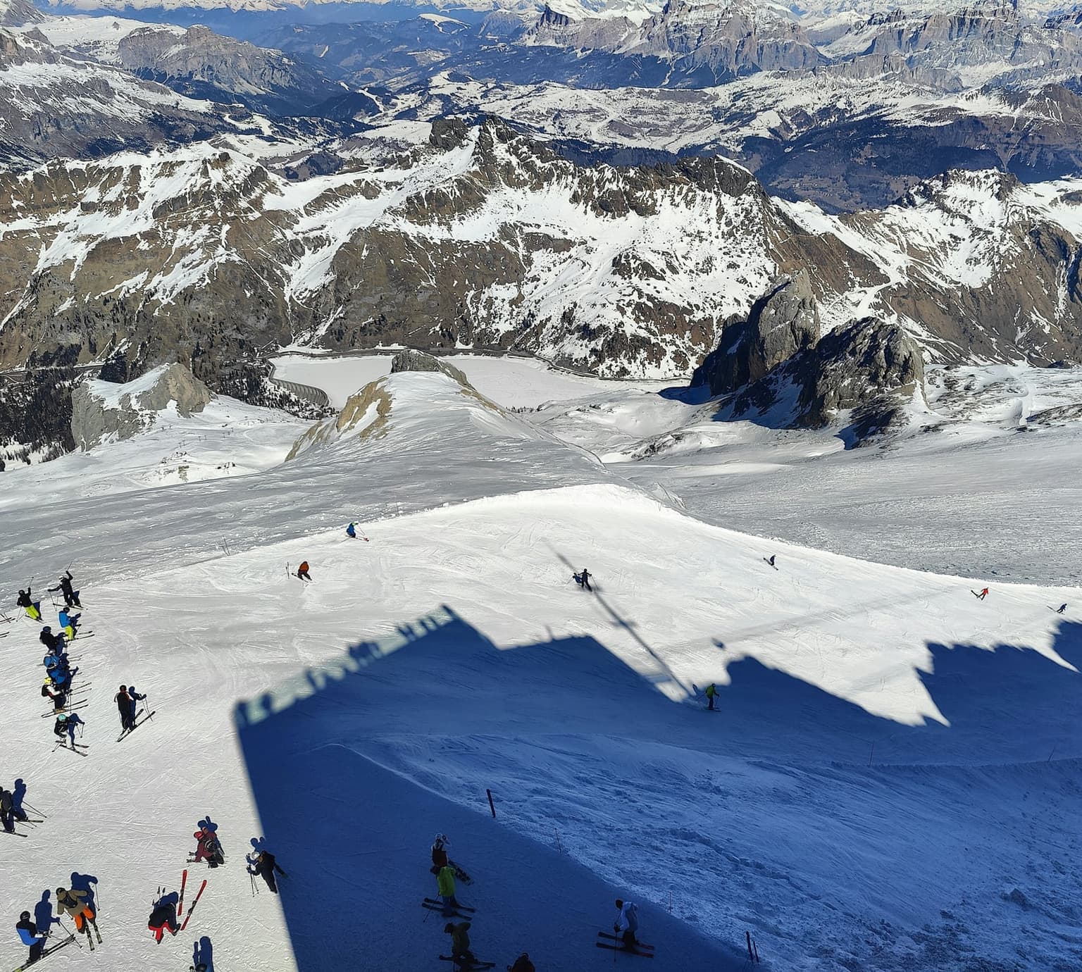 Looking down the Marmolada glacier piste from the top station, skiers at the start and others carving below, frozen Lake Fedaia and its dam in the valley, and rows of Dolomite peaks beyond