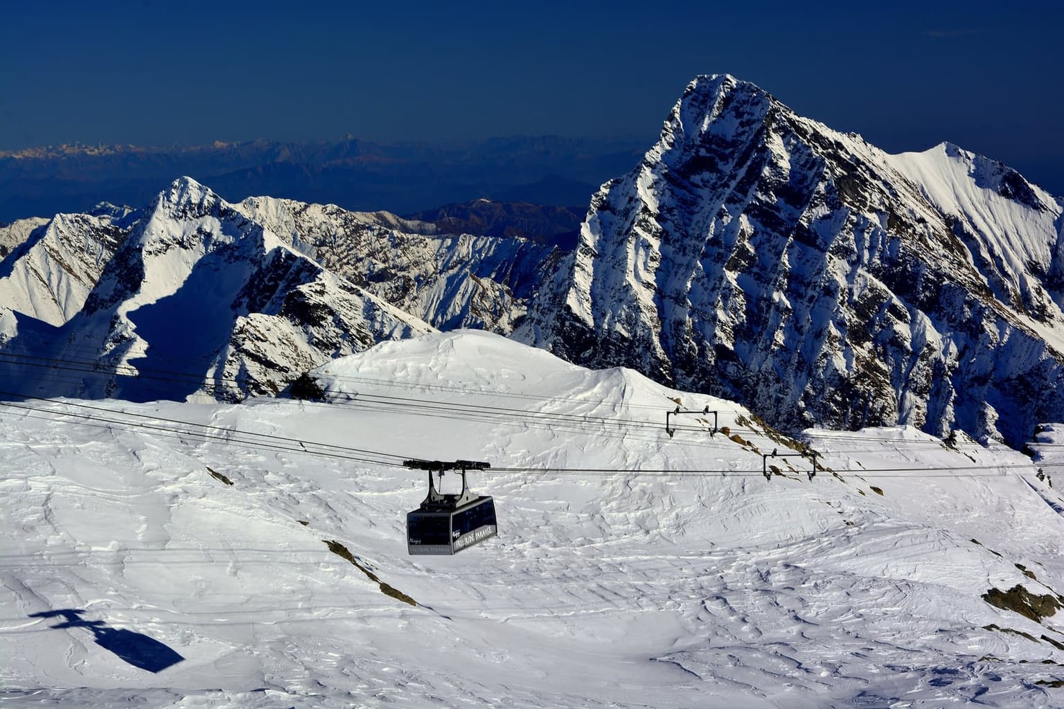 A large cable car cabin branded Alagna Free Ride Paradise crossing high, wind-sculpted snow slopes in the Monte Rosa area, with a steep snow-streaked rock peak behind it