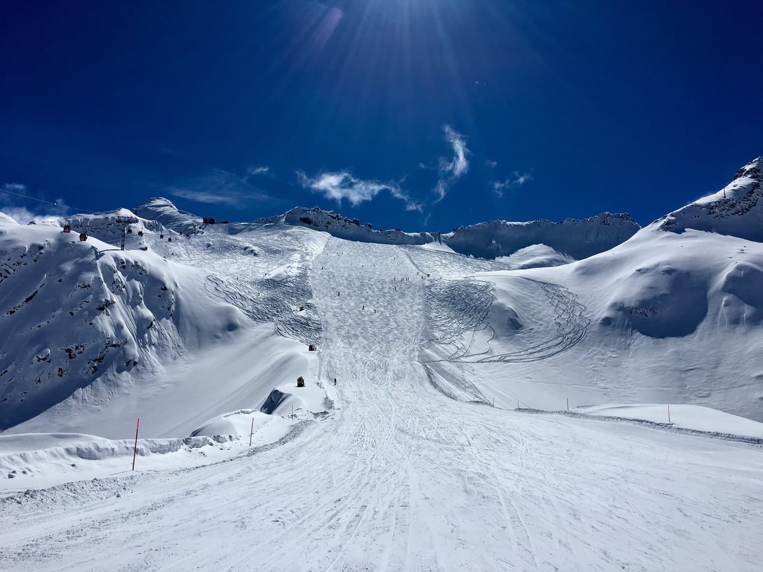 A wide, treeless glacier piste on the Presena above Passo del Tonale climbing toward a high snowy ridge, tiny skiers spread along it and a gondola line on the left