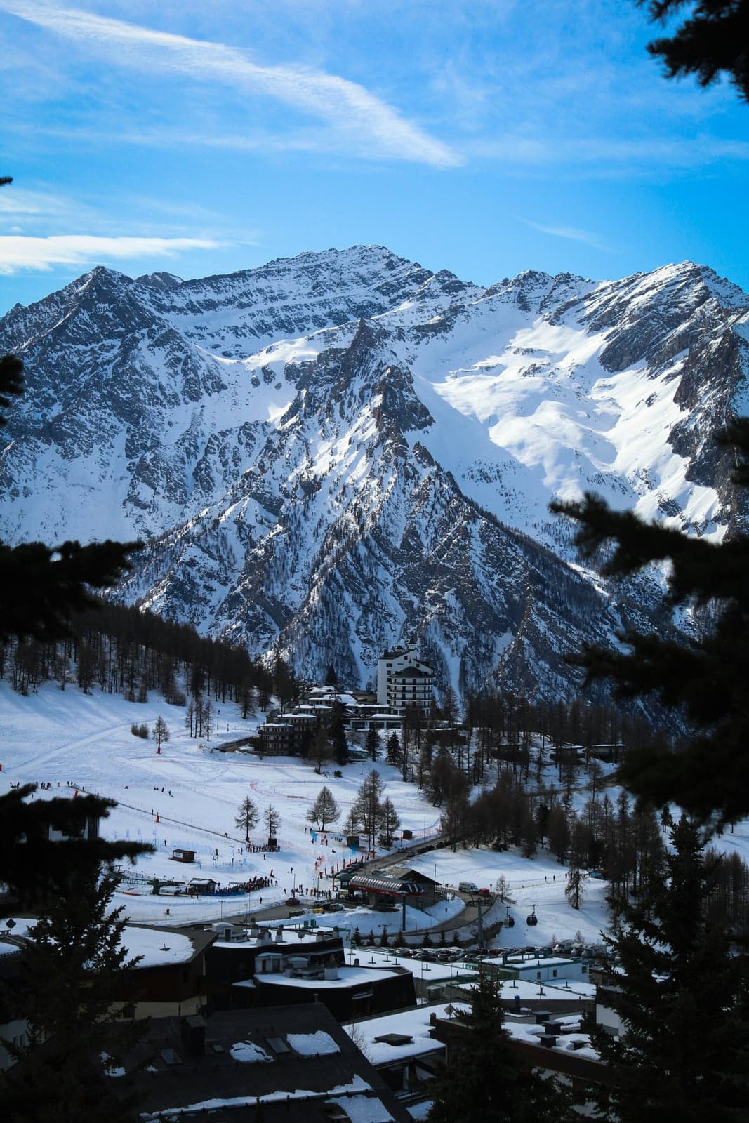 A ski resort village seen through dark conifers, with beginner slopes and a ski-school area, a tall white hotel and a gondola station below a large snow-covered mountain