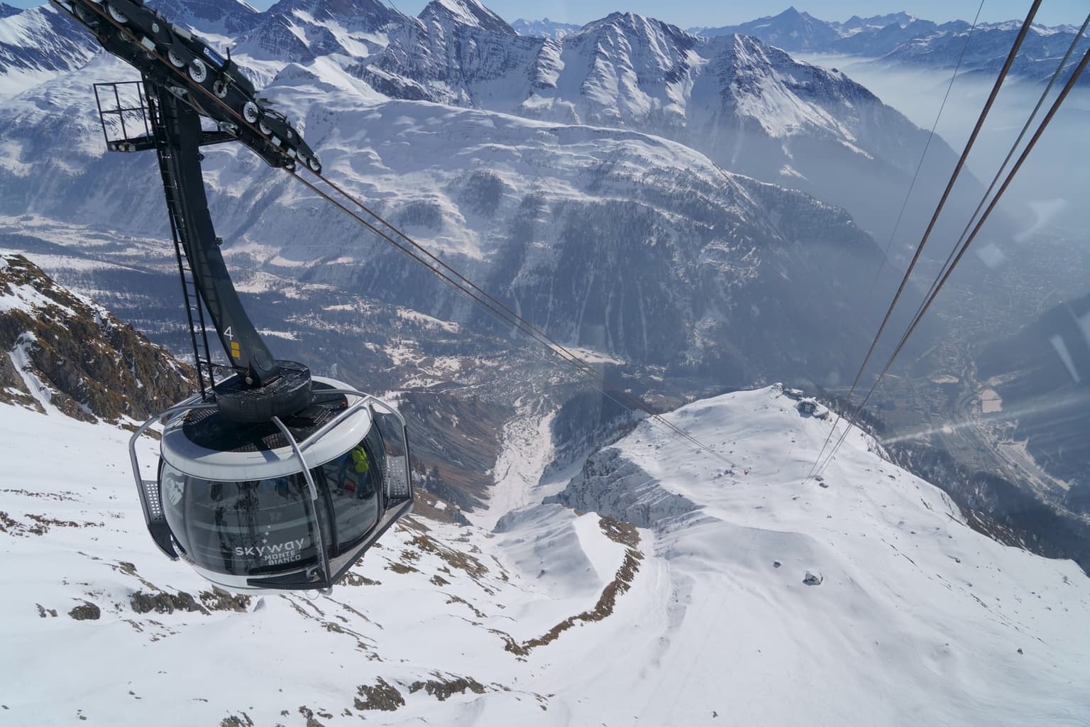 A round Skyway Monte Bianco cable car cabin passing a pylon high above Courmayeur, with snowy ridges, a station on a ridge below and the hazy valley far beneath
