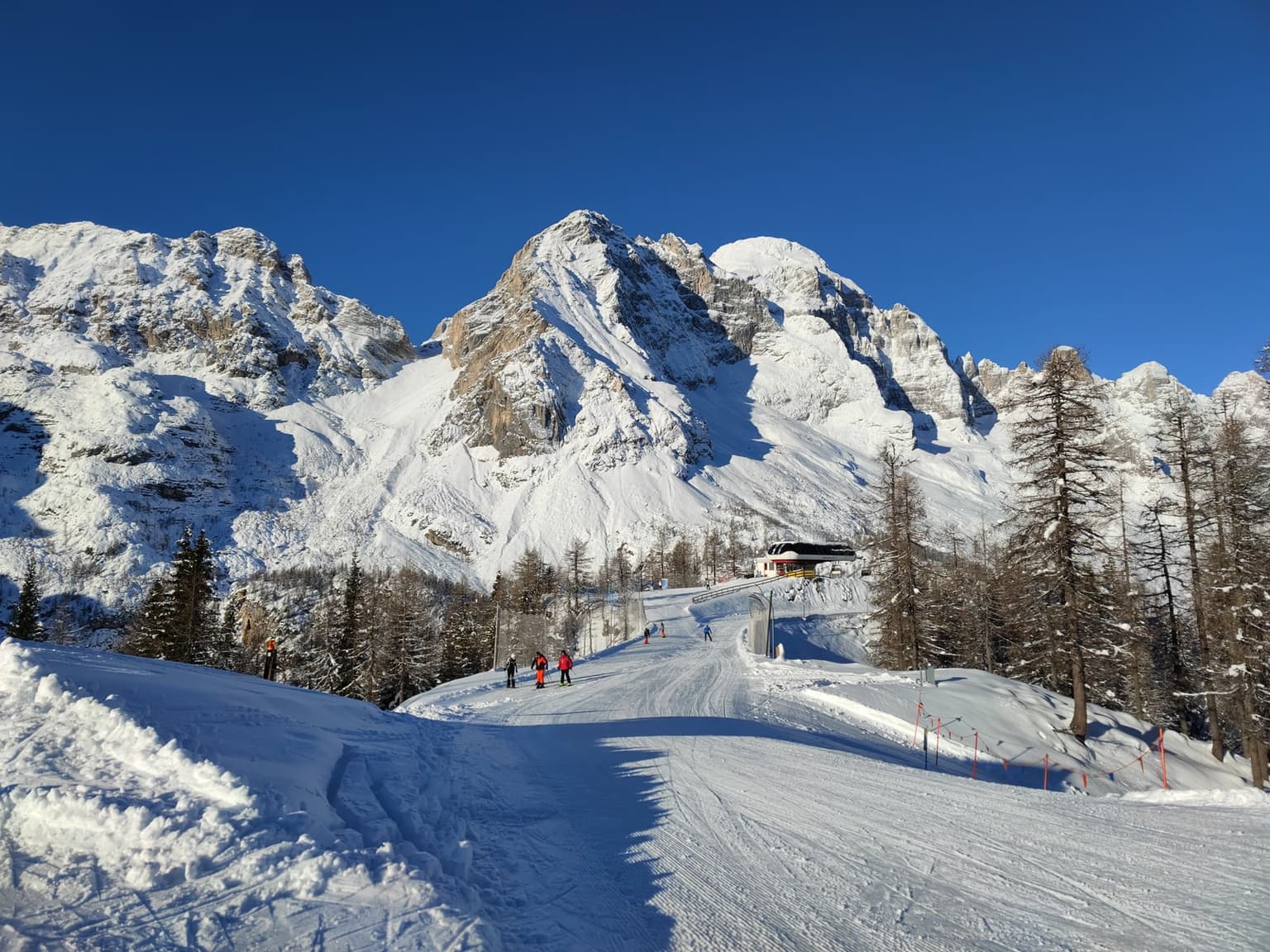 A wide groomed piste climbing between larches toward a chairlift station in the Dolomites, a few skiers on it and snow-covered rock walls rising behind under a deep blue sky