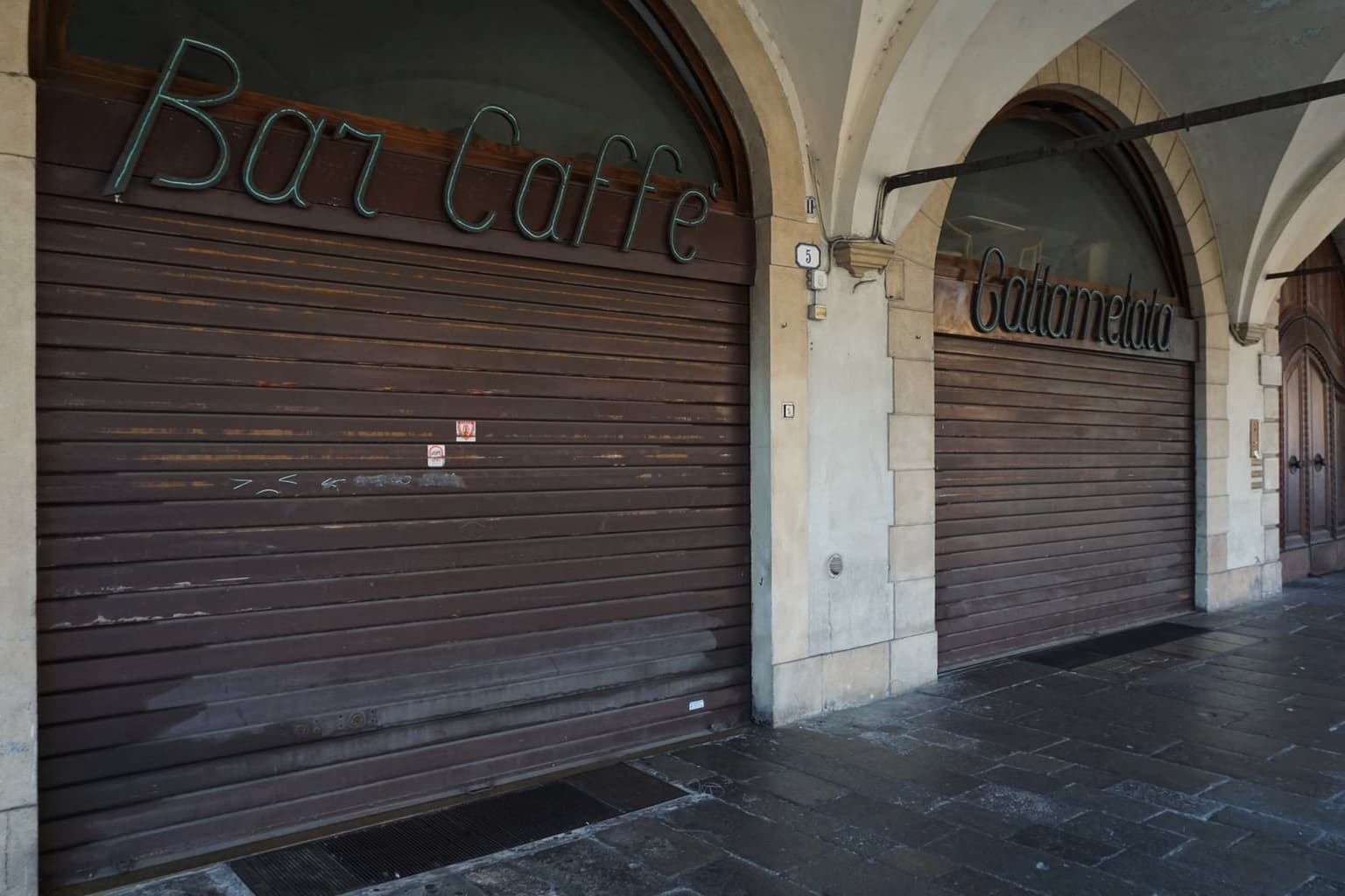 A shuttered Italian cafe under a stone archway, its roller shutter down beneath a neon-script "Bar Caffè" sign, a second closed storefront beside it on an empty arcaded sidewalk