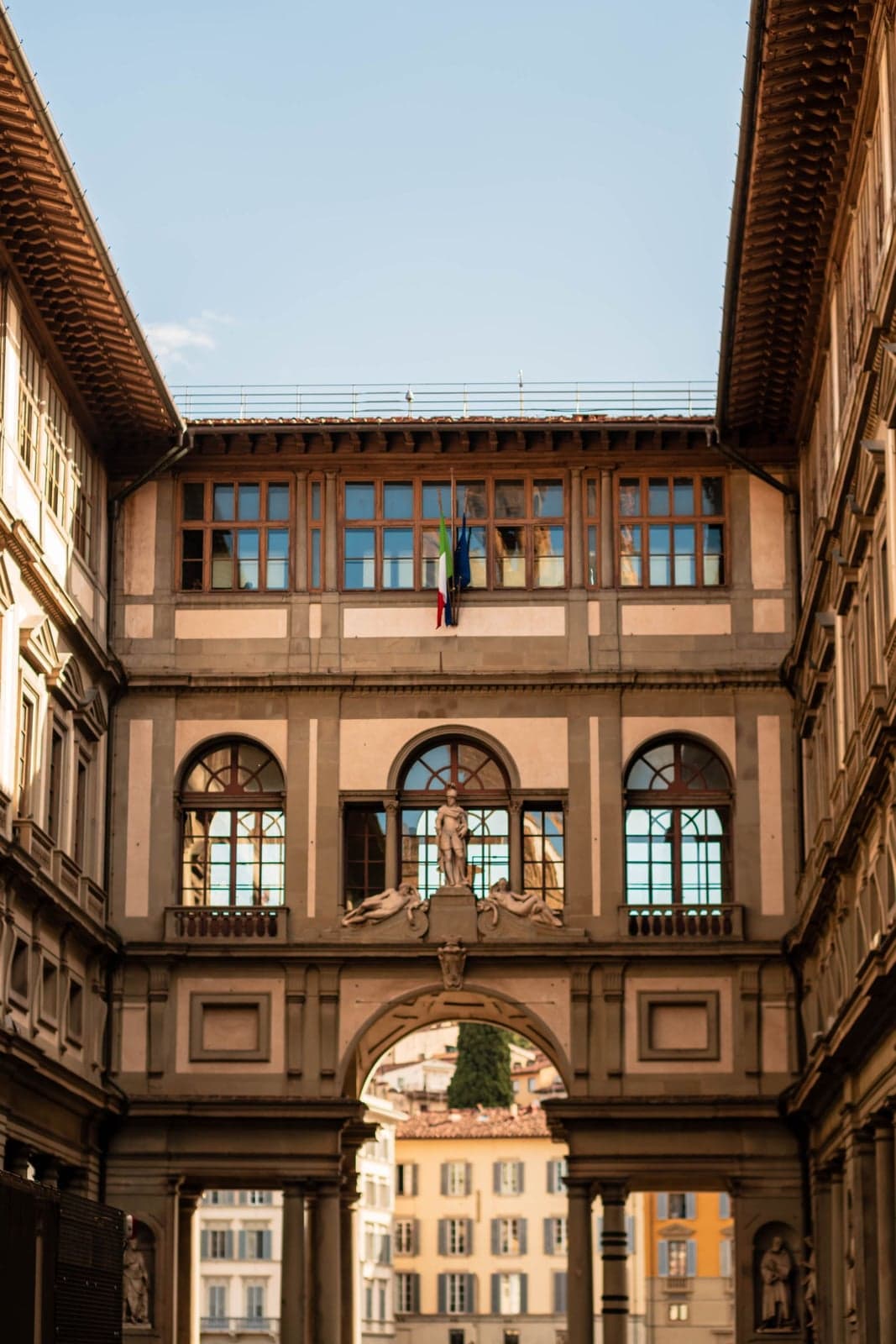 The narrow courtyard of the Uffizi Gallery in Florence, framed by matching wings of arched windows and statue niches, a triple-arched loggia opening onto a glimpse of the city's rooftops