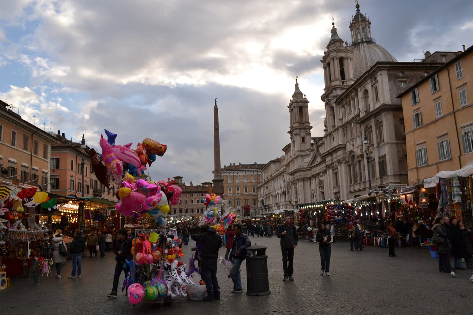 Piazza Navona in winter daylight, a Christmas market of stalls in the foreground, the obelisk and the dome of Sant'Agnese in Agone behind, pedestrians in dark coats crossing the square