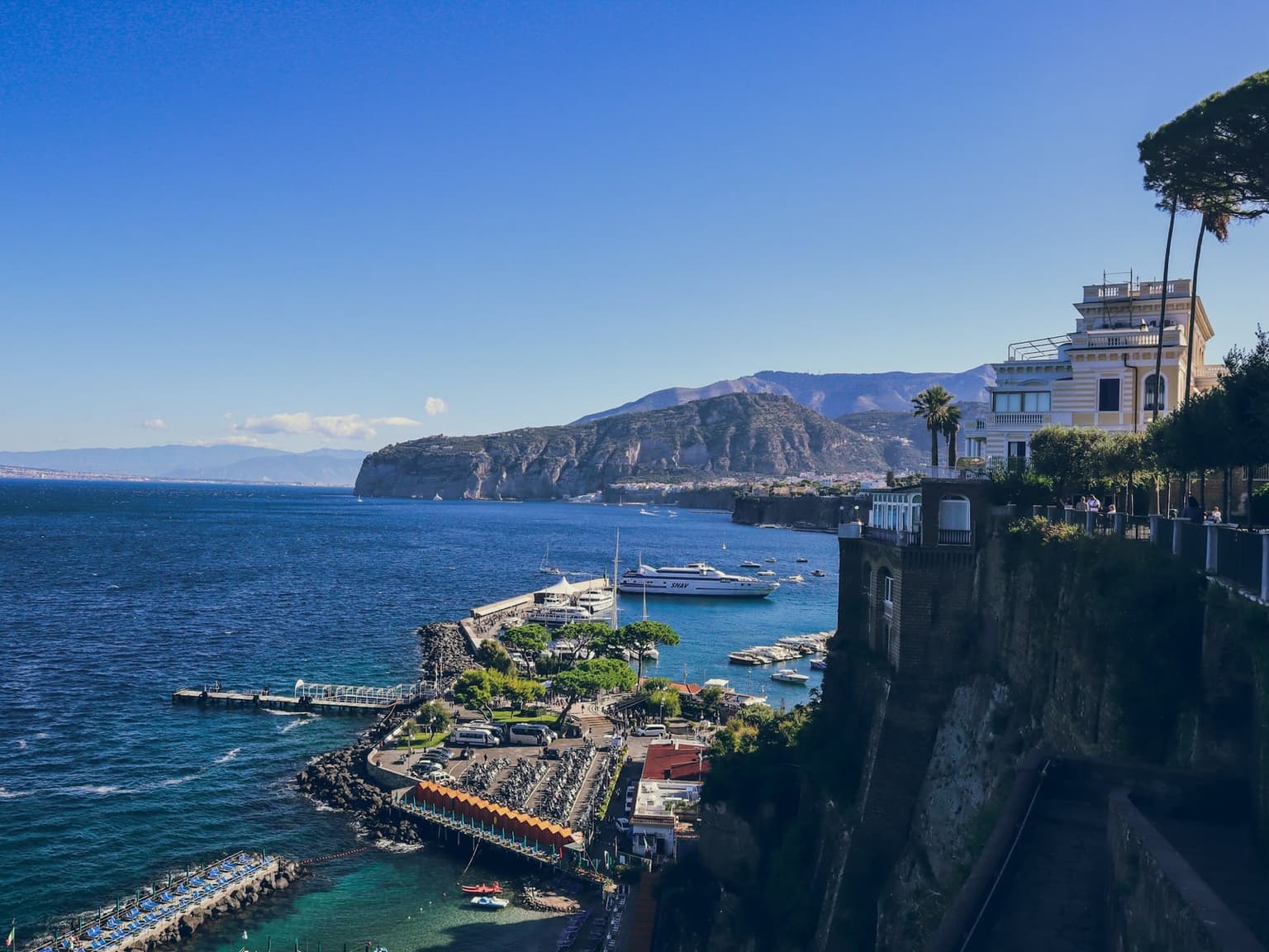 Marina Piccola, Sorrento's port at the foot of the cliff, seen from the clifftop, with a fast ferry at the pier, buses and parked scooters on the quay, beach-club platforms along the shore, and the coast curving east to a headland