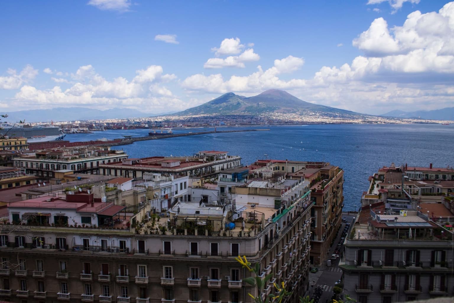 Naples from a hillside, rooftop terraces in front and the bay and harbor breakwater beyond, with a cruise ship docked at left and Vesuvius across the water