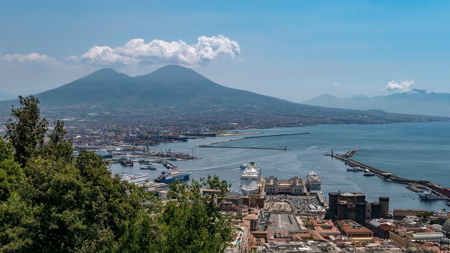 The port of Naples from a hillside above the city, with Castel Nuovo and the cruise terminal in front, ferries at the quays, a fast boat leaving between the breakwaters, and Vesuvius across the bay