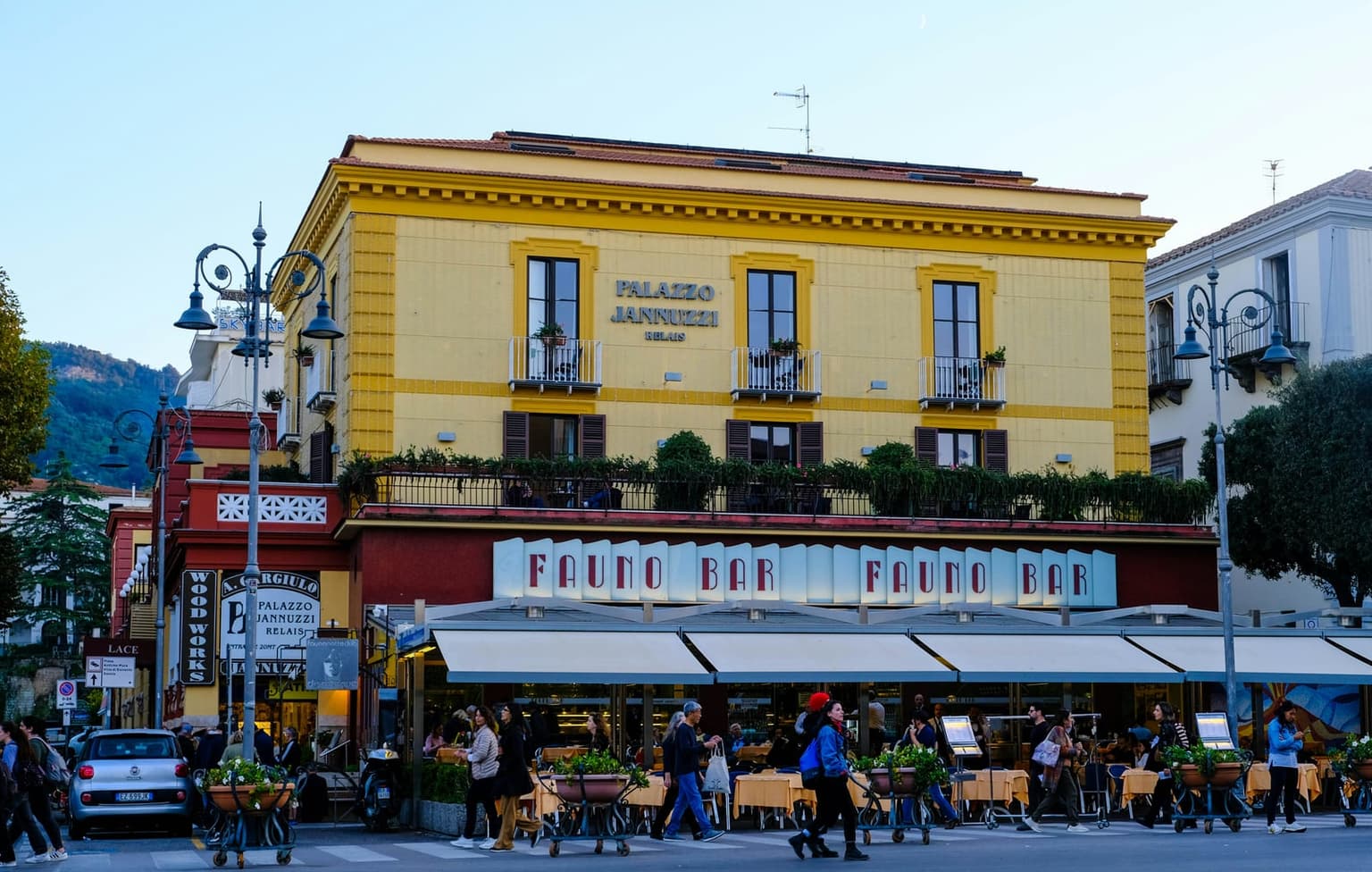 The corner of Piazza Tasso in Sorrento in early evening light: the yellow Palazzo Jannuzzi above the Fauno Bar's café terrace and white awnings, with people crossing the square