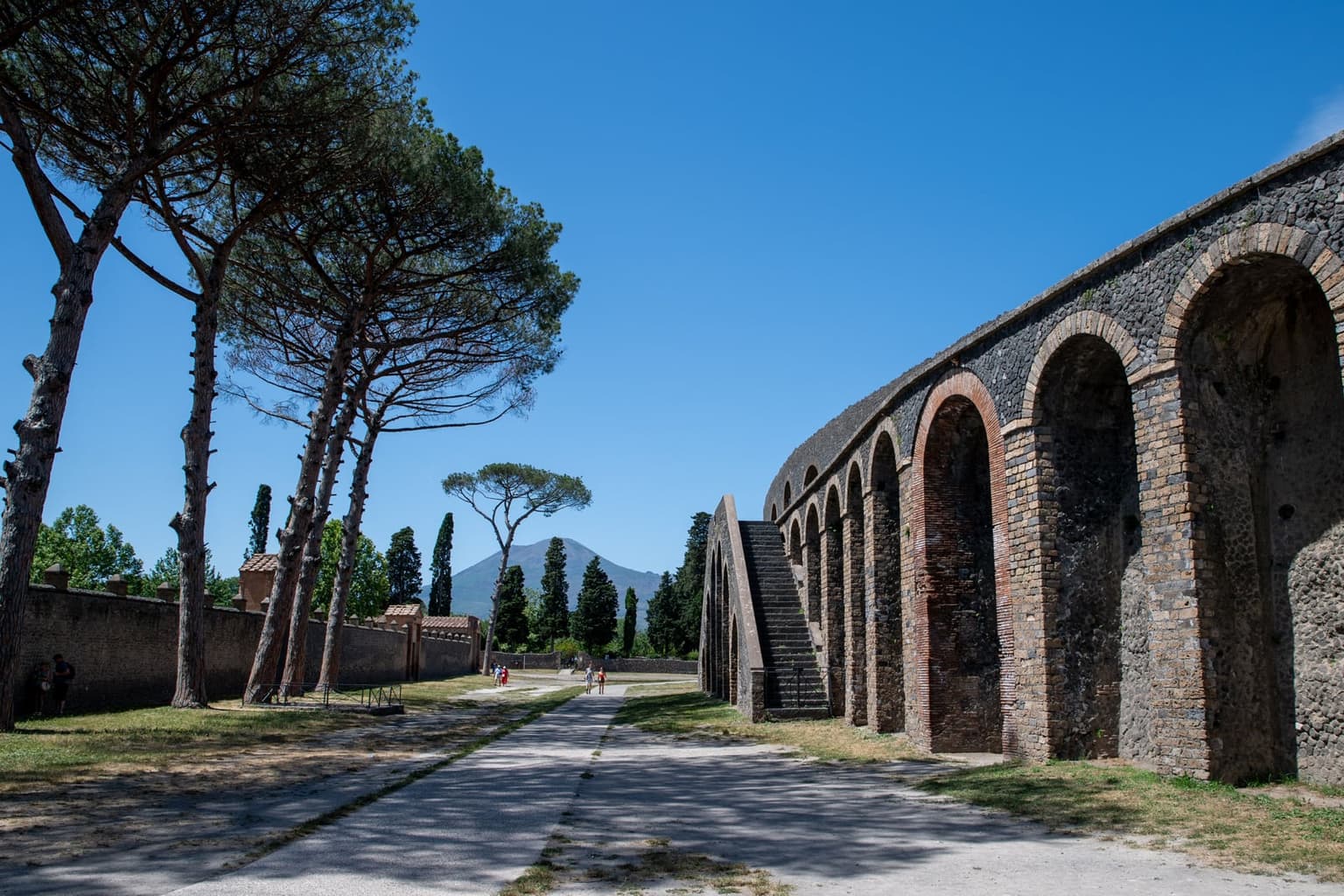 The arched outer wall and stone staircase of Pompeii's amphitheater beside a path lined with umbrella pines, with Vesuvius at the end of the path under a clear blue sky