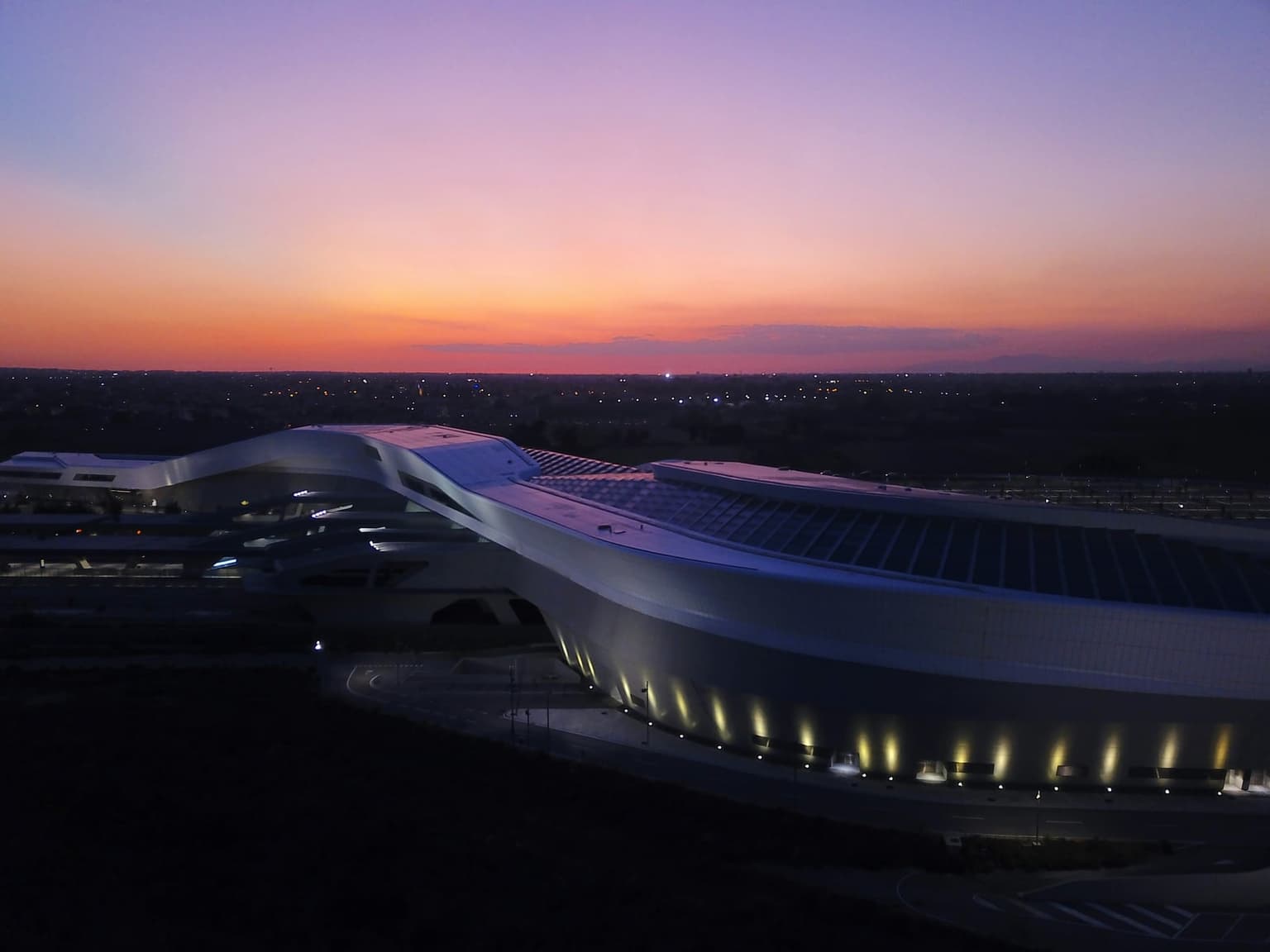 Napoli Afragola high-speed station from above at dusk: the long, curving white station building with its ribbed glass roof, lit along its base under an orange and violet sky