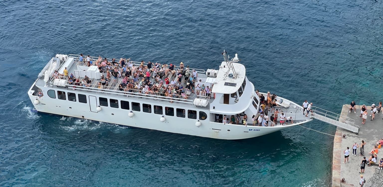 A white passenger ferry with a crowded open upper deck and a gangway out to the small stone quay at Vernazza, seen from above as people board and others wait on the quay.