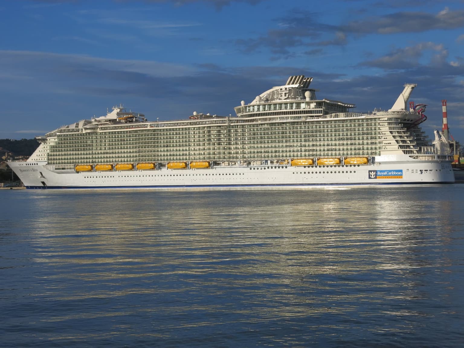 A very large white cruise ship berthed in La Spezia's port in low evening light, with a row of yellow lifeboats along its hull and a cloudy blue sky above calm water.