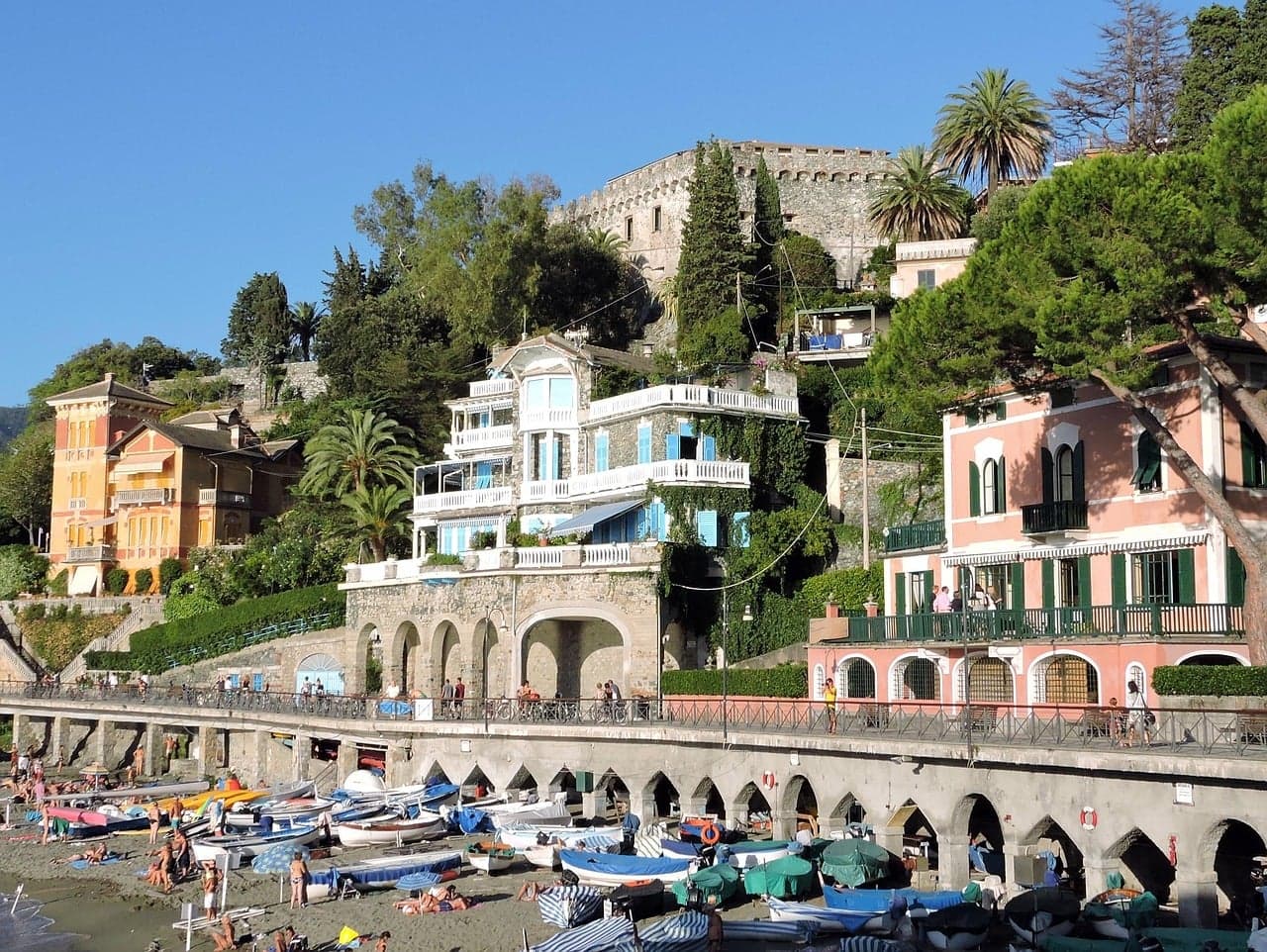 Levanto's arcaded sea wall and promenade above a beach lined with small boats, with pastel villas, palm trees and the castle wall on the hill behind.