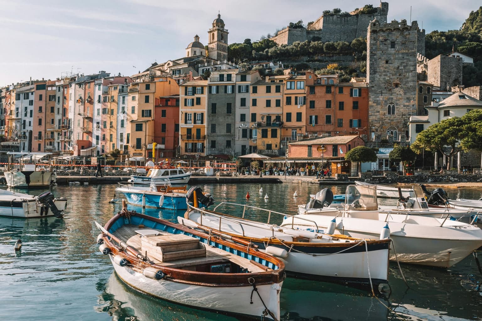 The tall pastel harbor front of Portovenere with a crenelated stone tower, a church bell tower and the hilltop castle above, seen across still water past moored wooden boats.