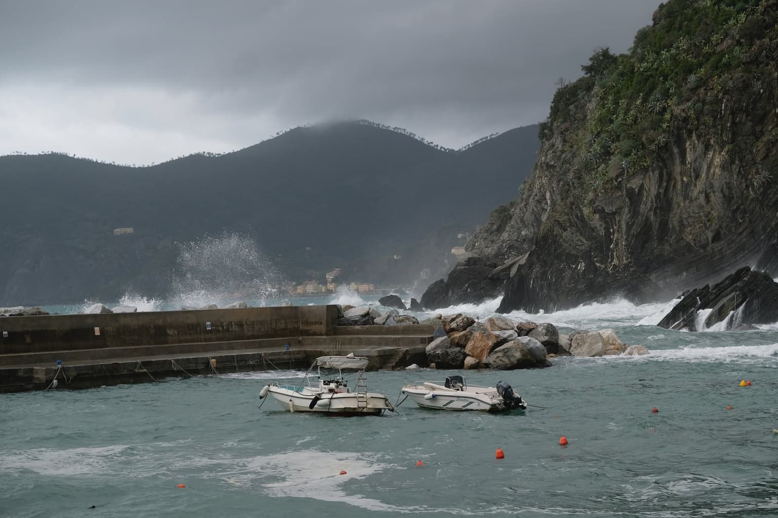 Two small boats ride out rough green water inside Vernazza's harbor while waves break over the breakwater and a steep rocky cliff, under dark storm cloud on the hills behind.