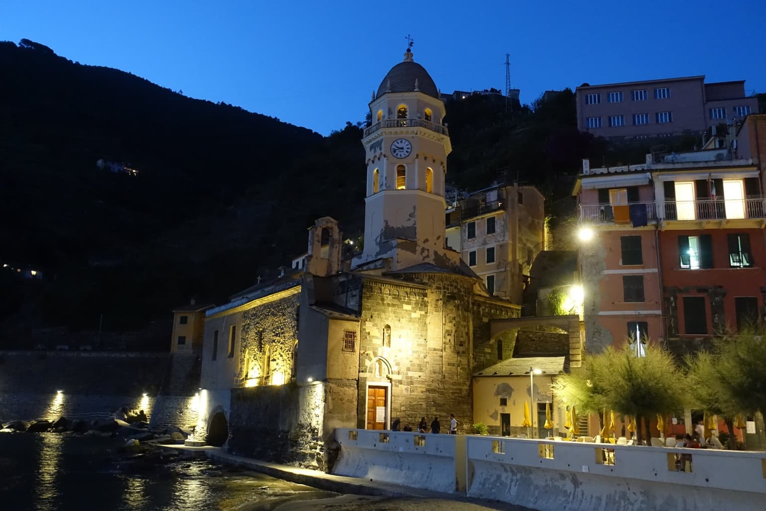 Vernazza's lit bell tower and stone church at blue hour, with glowing windows in the pastel houses beside them, dark water below and a few people along the sea wall.