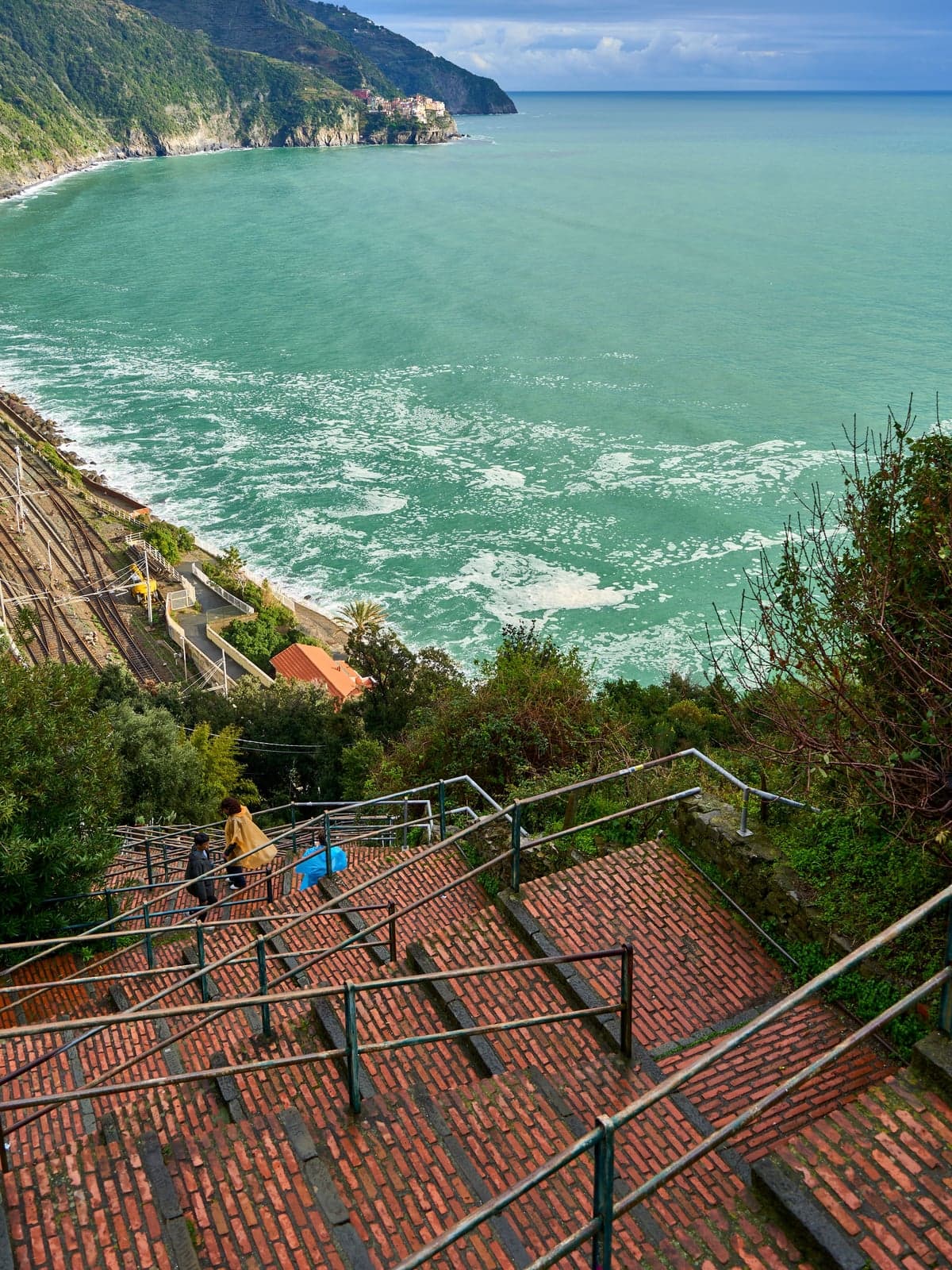Corniglia's brick Lardarina stair zigzagging down a steep hillside toward the railway and a turquoise sea, with two walkers in rain ponchos on the steps and a village on a far headland.
