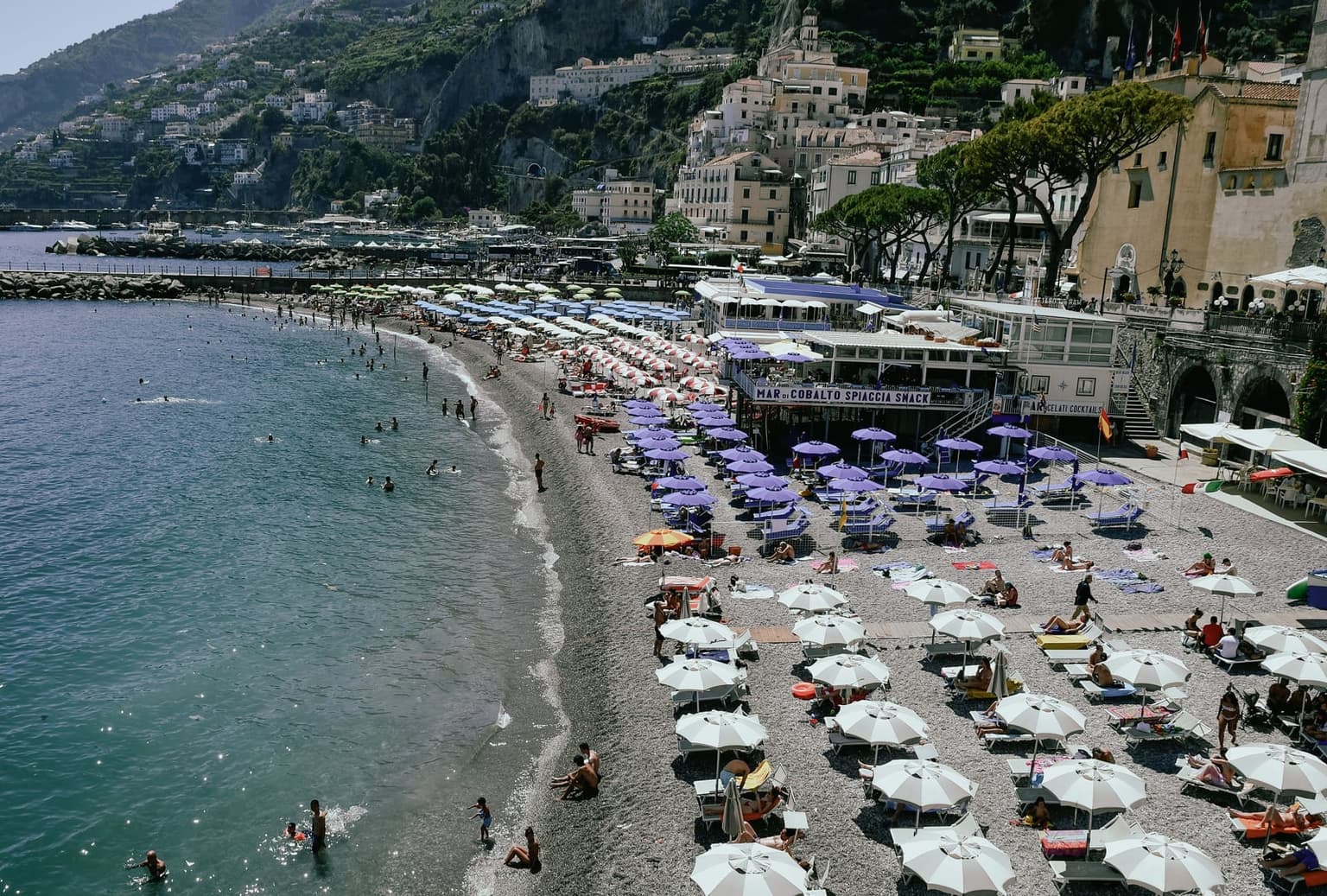 Amalfi's town beach from above, gray pebbles almost entirely covered by rows of paid loungers under white, purple, and pink umbrellas, swimmers at the water's edge