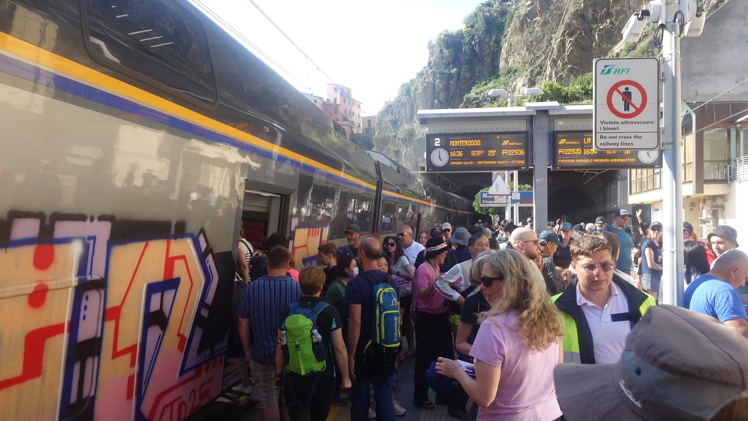 A dense crowd of day-trippers with backpacks pushing to board a regional train at Manarola station, the departure board showing a delayed train to Monterosso and a railway tunnel behind