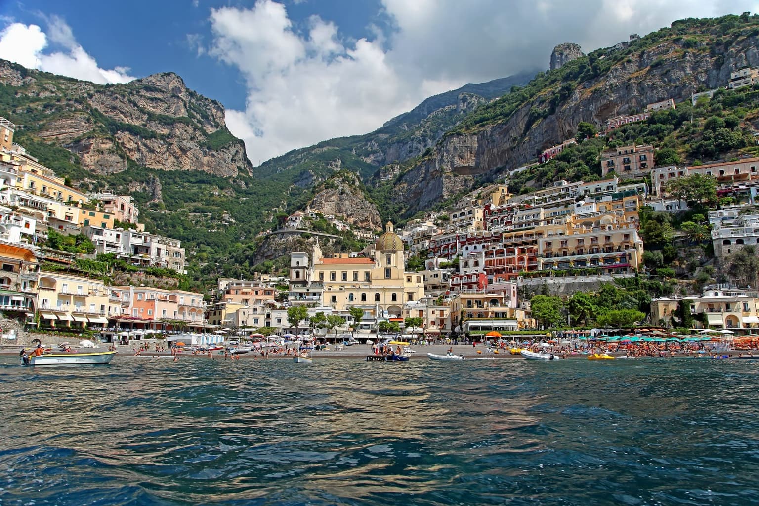 Positano from the sea, pastel houses stacked straight up the mountainside from a crowded beach, the tiled dome of Santa Maria Assunta at the waterfront and sheer limestone cliffs above