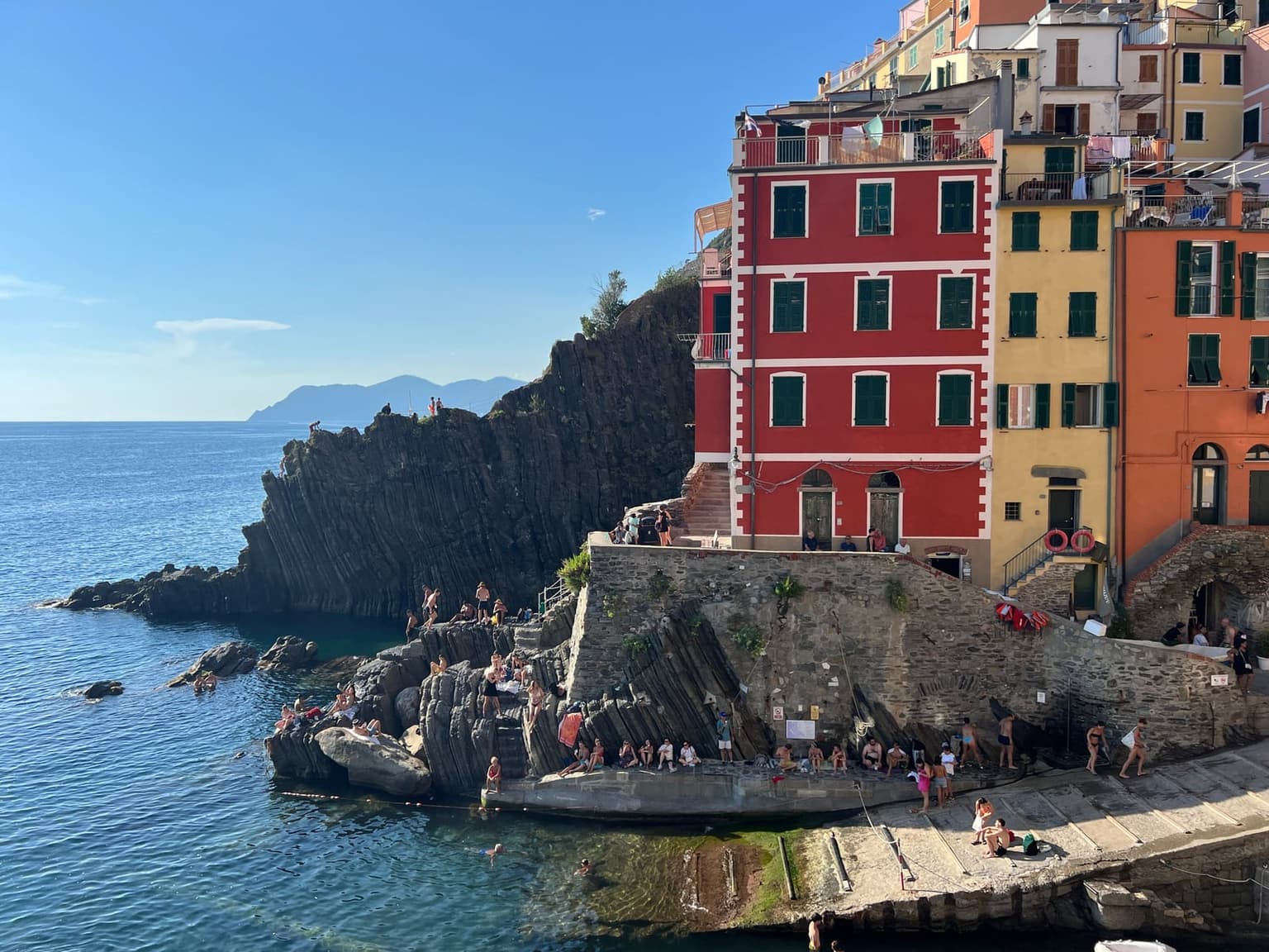 Swimmers in clear water at Riomaggiore's small harbor, sunbathers on tilted dark rock ledges and along the stone quay below a red house with green shutters