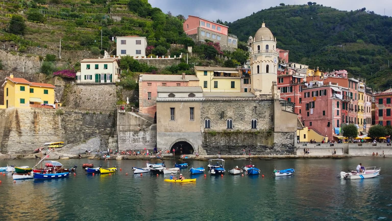 Vernazza from the water, small boats and a yellow kayak moored in the harbor below the stone church with its domed bell tower, pastel houses and terraced vineyards stacked up the hillside behind