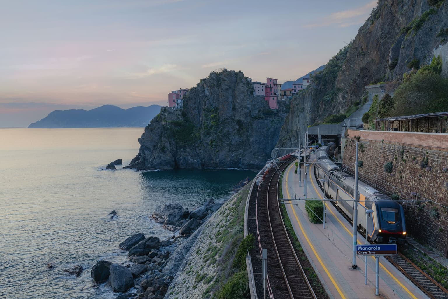 A regional train arriving at Manarola's clifftop station at dusk, the coastal track hugging the rock face above a small cove with the sea directly beside the platform.