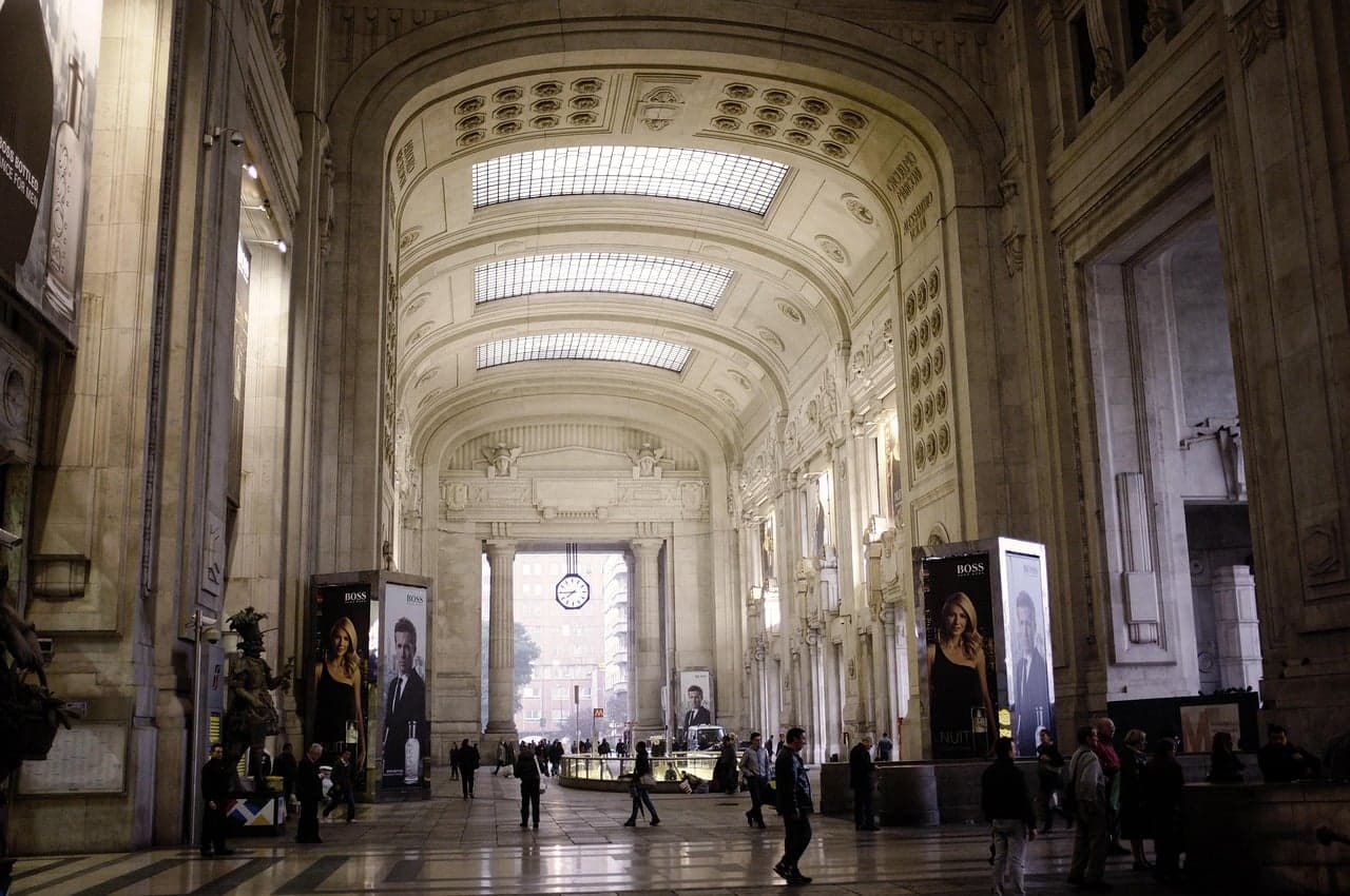 The vaulted departures hall at Milano Centrale, its arched glass skylights and columned galleria leading toward a large station clock and the street beyond.