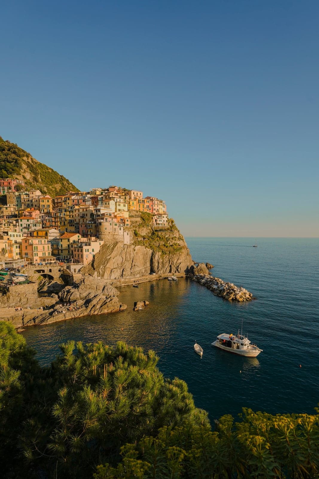 A motorboat and a small dinghy anchored in Manarola's harbor, the village's pastel cliffside houses rising above the water and a stone breakwater curving out into the sea.