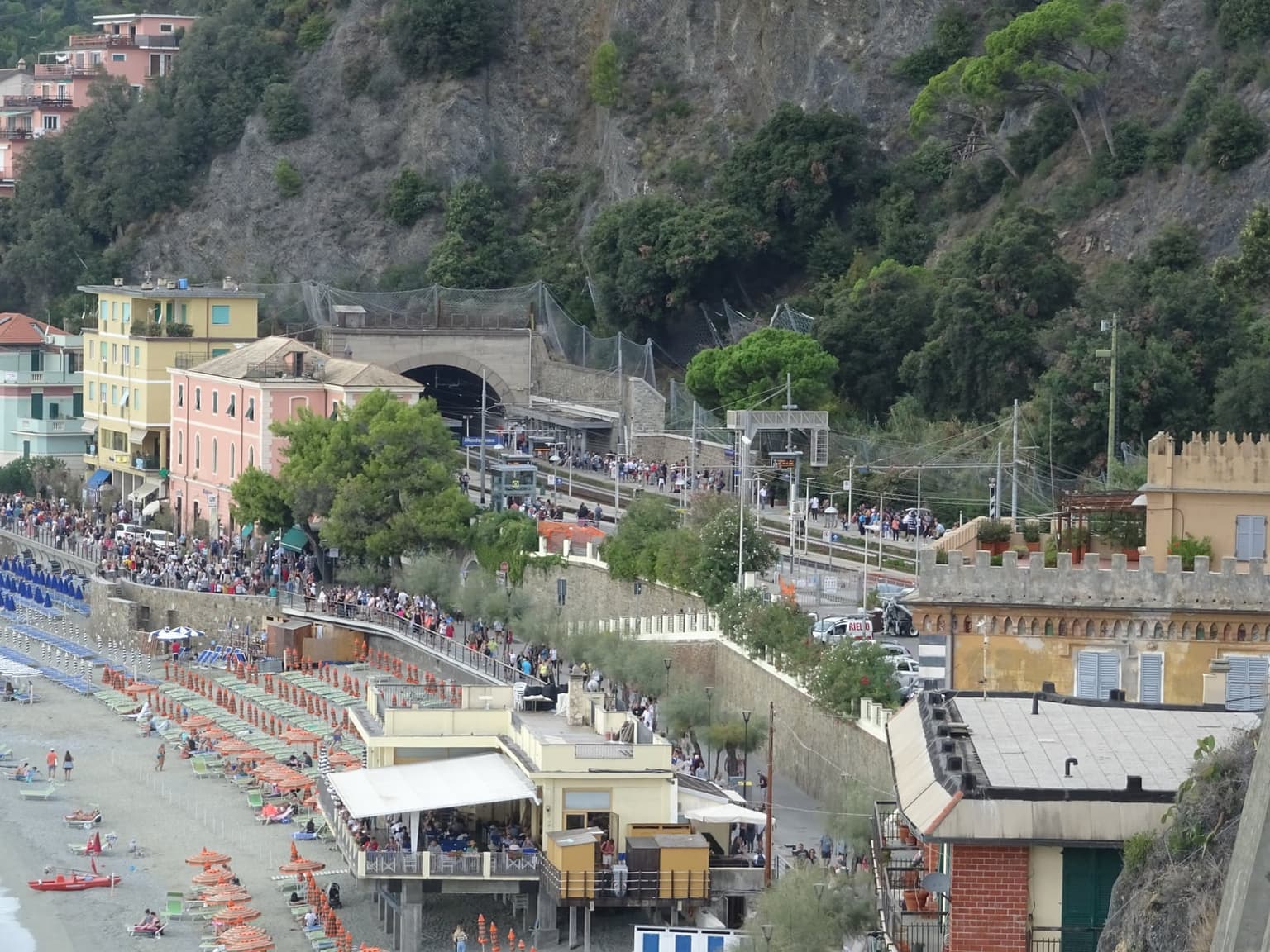 Elevated view of Monterosso al Mare's railway station on the Fegina seafront, with the platform and a hillside rail tunnel running alongside a crowded beach lined with orange umbrellas, the sea just beyond.