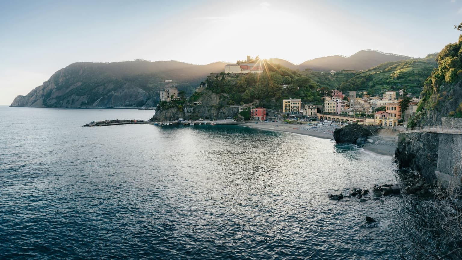 Monterosso al Mare in late-afternoon light, seen from the cliff path above the sea, its old town and arched railway viaduct behind a curved beach and the low sun behind the convent hill