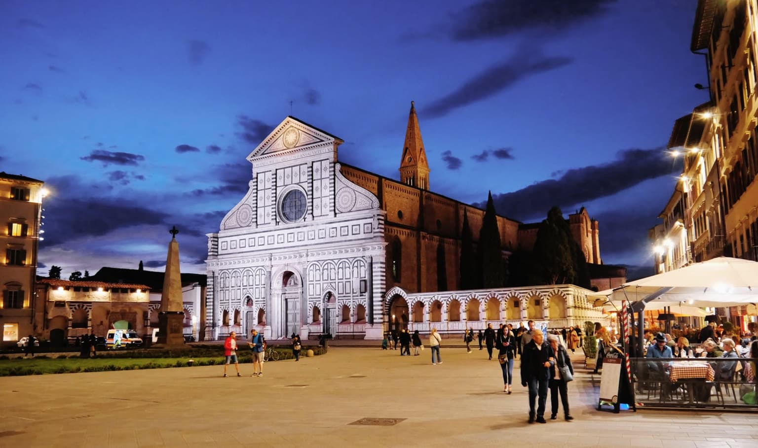 The white-and-green marble facade of the Basilica di Santa Maria Novella in Florence, floodlit at dusk, with people strolling across the piazza and restaurant tables under white umbrellas on the right
