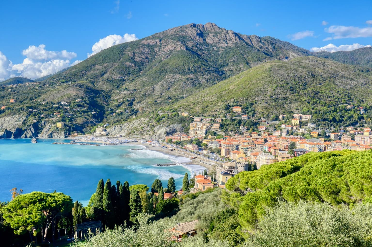 Levanto from the hillside above town, its orange-roofed houses spread behind a long curved beach, with a small harbor at the far end of the bay and a green mountain rising behind