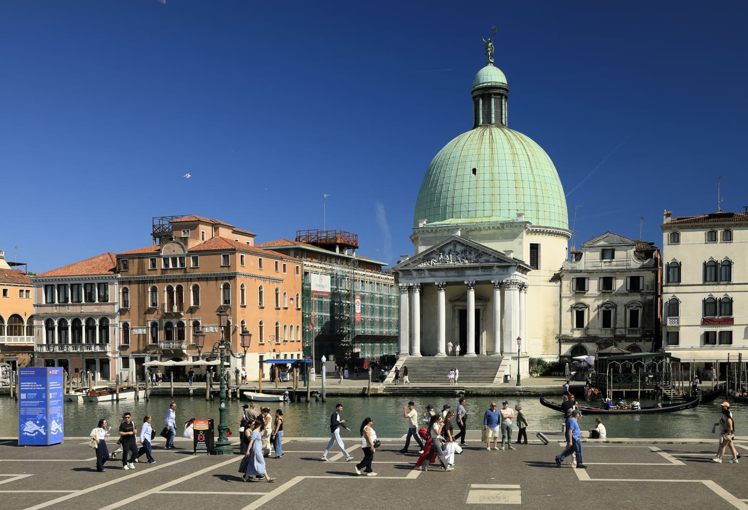 The green dome of San Simeon Piccolo across the Grand Canal, seen from the forecourt outside Venezia Santa Lucia station, with people crossing the paving, one pulling a wheeled suitcase