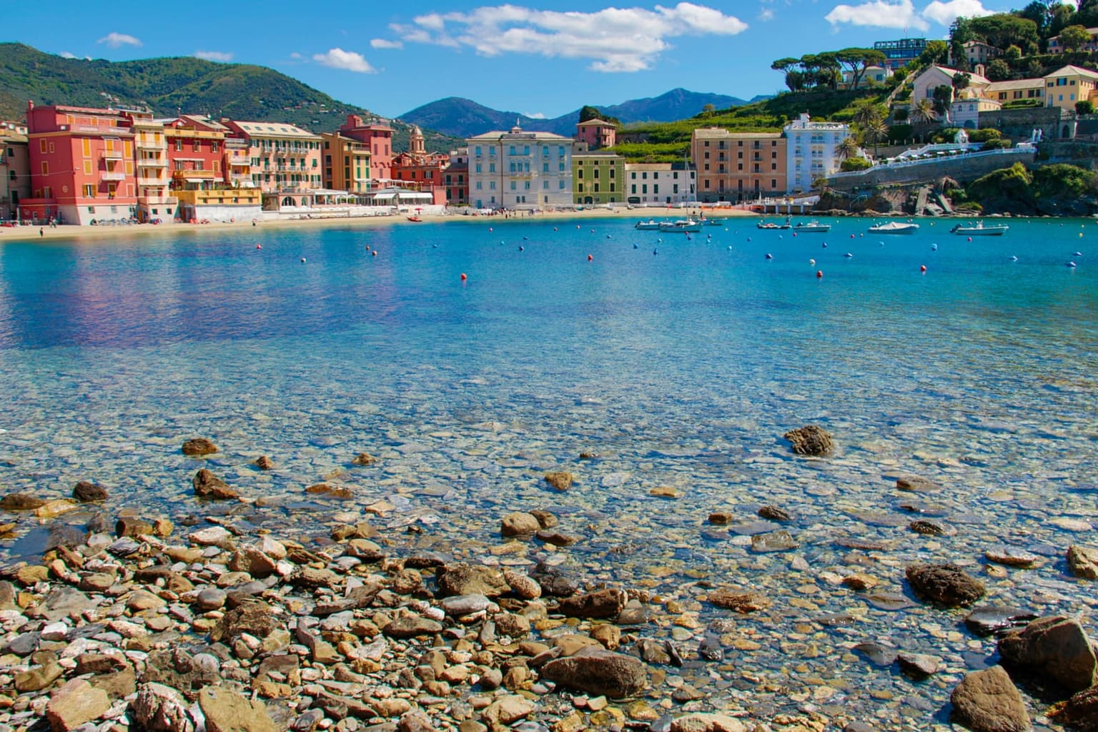 Sestri Levante's Bay of Silence, a curve of tall red, orange and yellow houses above a narrow beach, with small boats on clear turquoise water and wooded hills behind