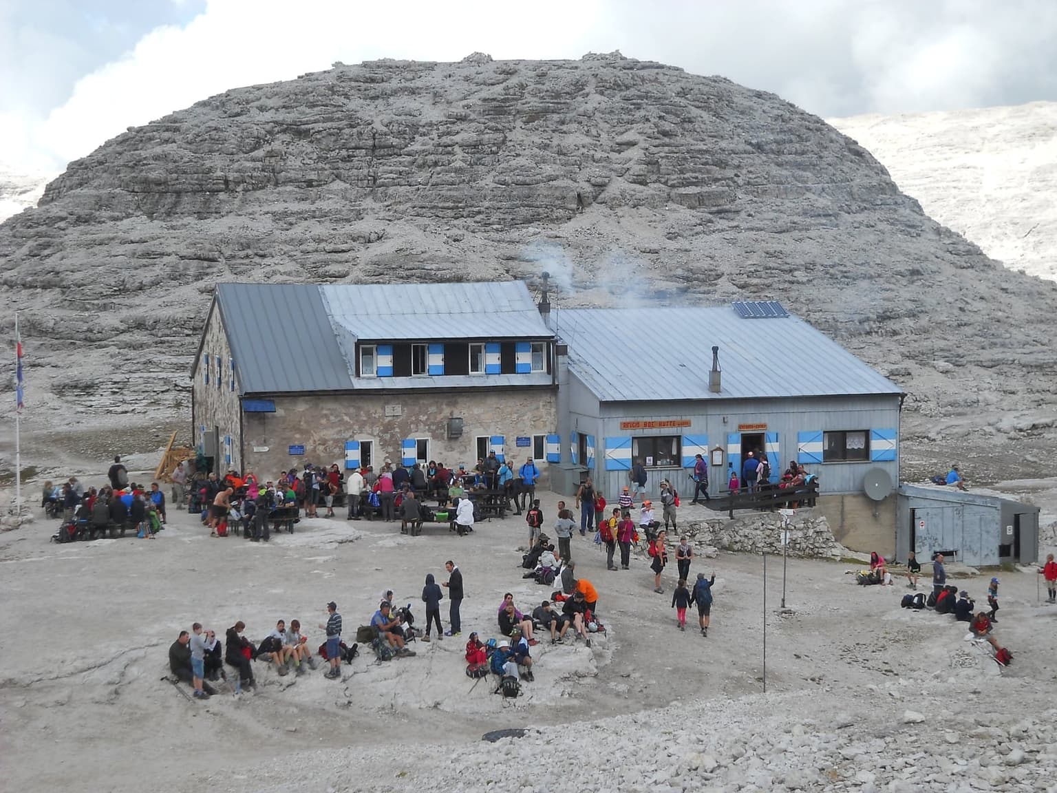 Rifugio Boè, a stone-and-timber hut with blue-and-white shutters, on the bare gray limestone of the Sella plateau, hikers crowding its outdoor tables and the rock in front