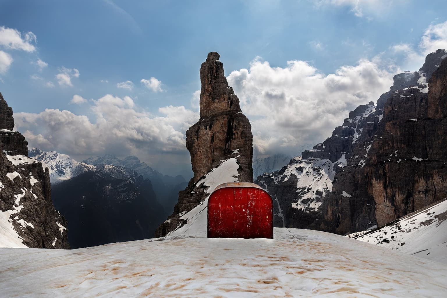 A small red half-barrel metal bivouac shelter on spring snow at the foot of the Campanile di Val Montanaia, a lone rock spire between snowy Dolomite walls