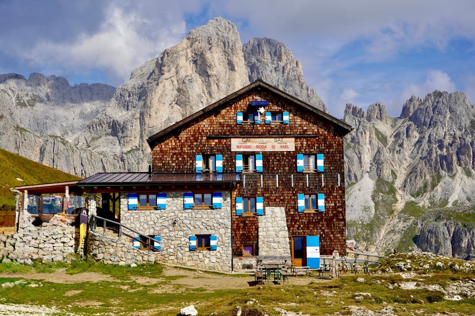 Rifugio Roda di Vael, a shingled wooden hut on a stone ground floor with blue-and-white shutters and its CAI SAT sign on the facade, picnic tables on the grass in front and pale, jagged Dolomite walls rising behind