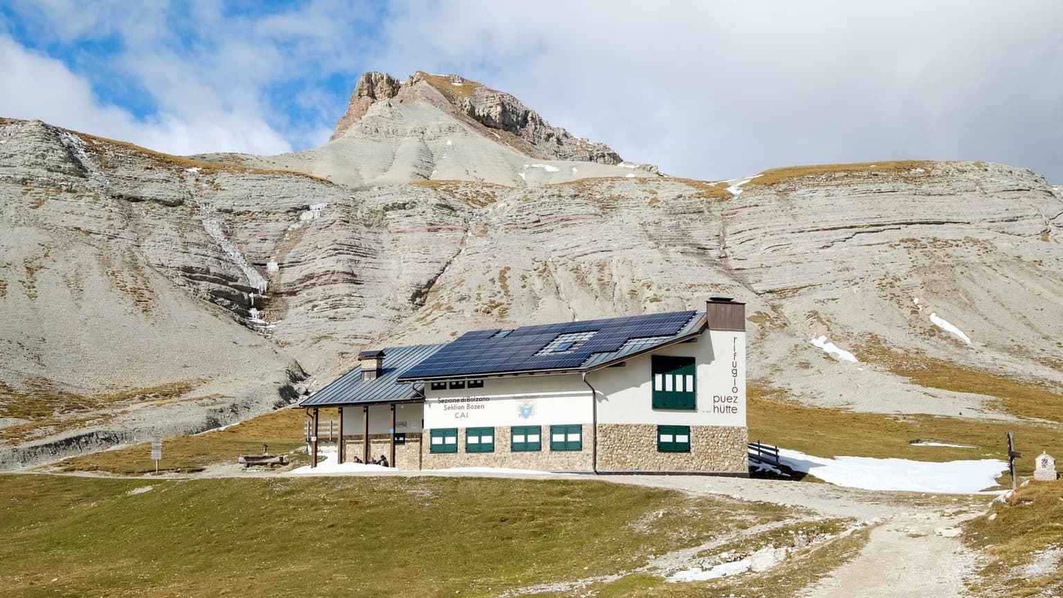 Rifugio Puez, a white hut on a stone base, on the autumn-brown grass of the Puez plateau, banded gray scree slopes rising behind and patches of old snow by the path